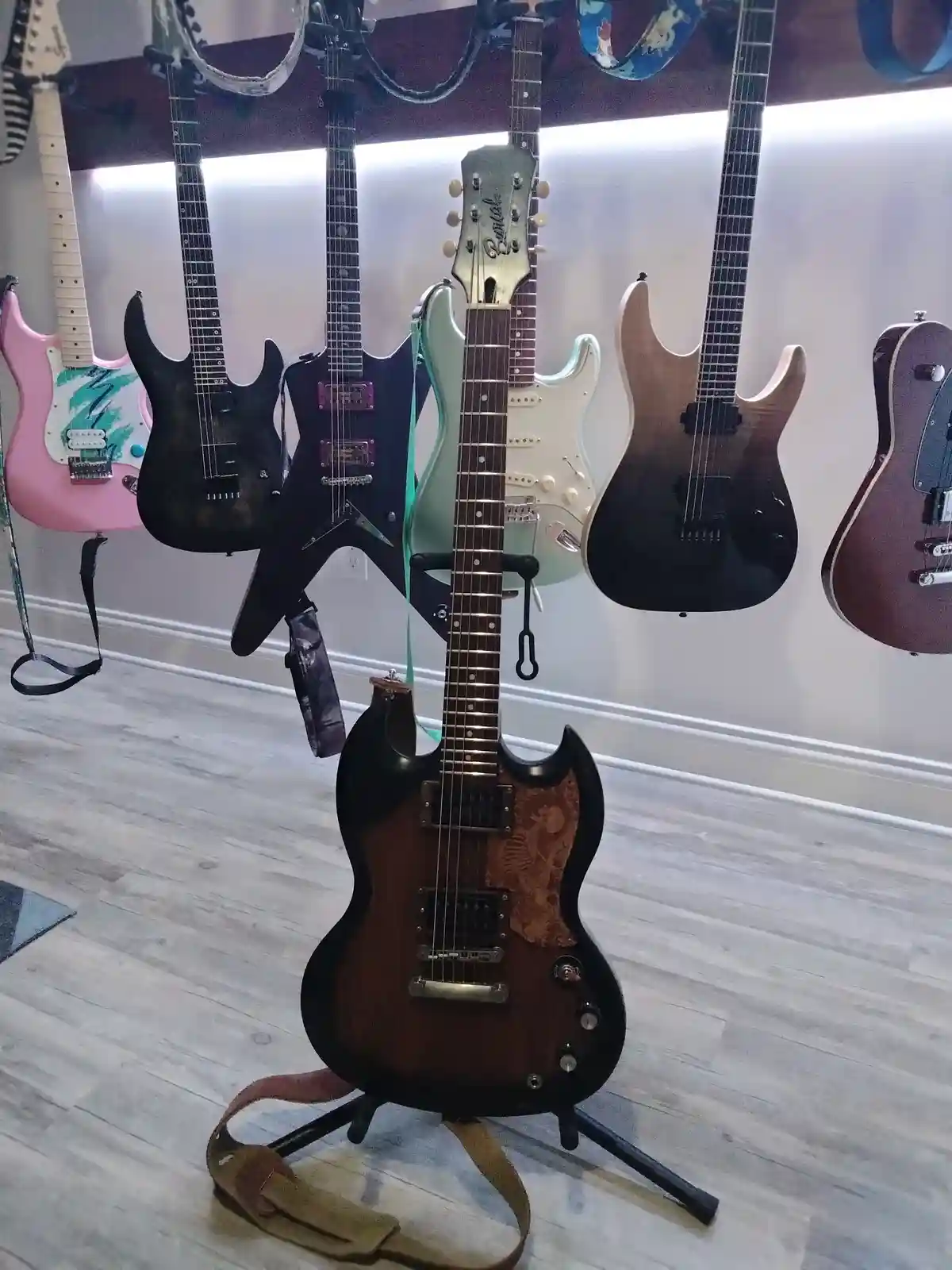 A brown electric guitar with ornate inlay sits on a stand in the foreground, with several other guitars hanging on a wall…