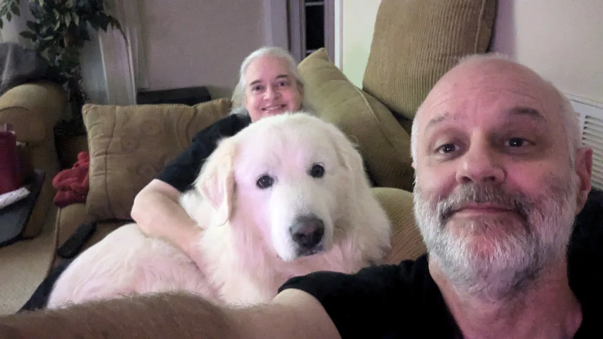 A selfie-style photo shows a bald man with a gray beard taking a picture with a large white dog and a woman with long gray…