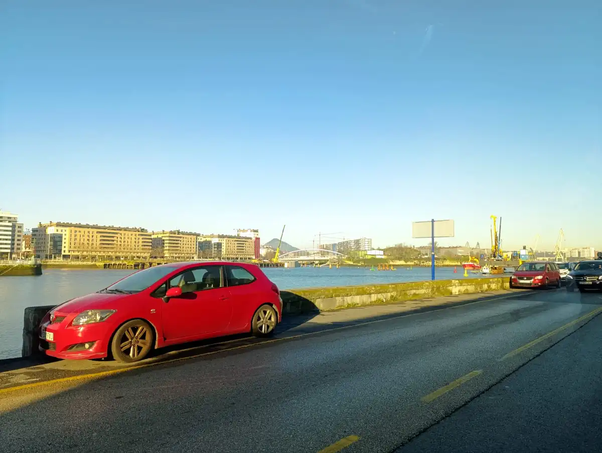 A red car is parked on the side of a road next to a body of water with a city skyline in the background. The overlaid text…
