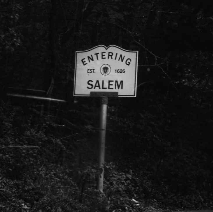 A black-and-white photo shows a roadside sign that reads 'ENTERING EST. 1626 SALEM' mounted on a post in a wooded area. The…