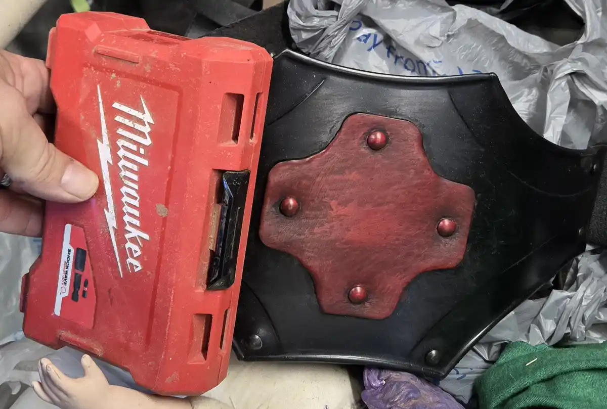 A hand holds a red Milwaukee Shockwave battery pack next to a black cosplay armor piece with a red, riveted plate, creating…
