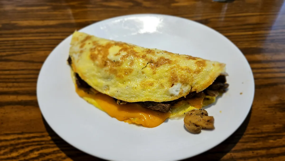 A close-up photo shows a folded, golden-brown omelette on a white plate, resting on a wooden table. Melted orange cheese and…
