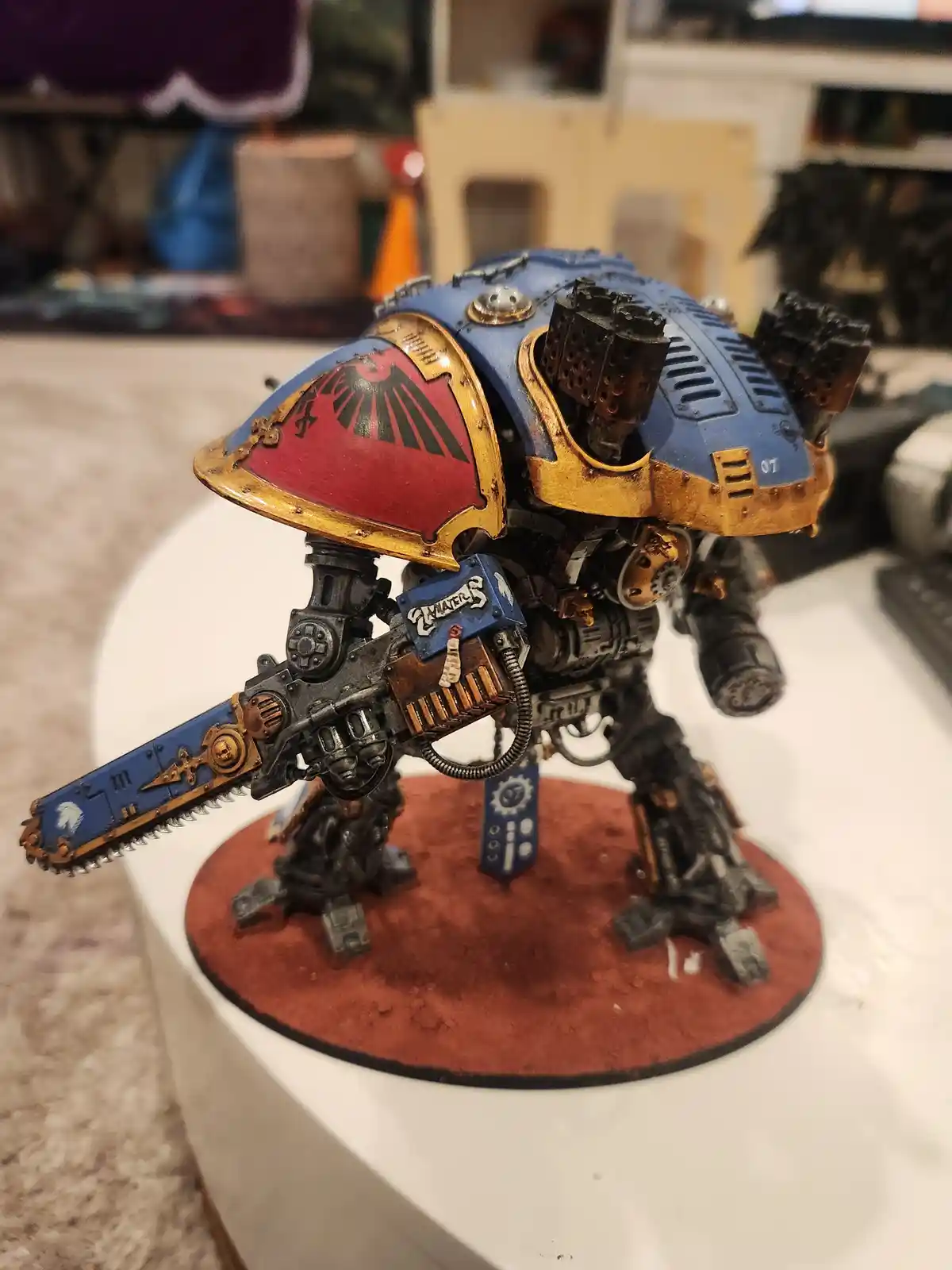 A highly detailed, hand-painted miniature of a blue and gold armored space marine wielding a chainsword, standing on a red…