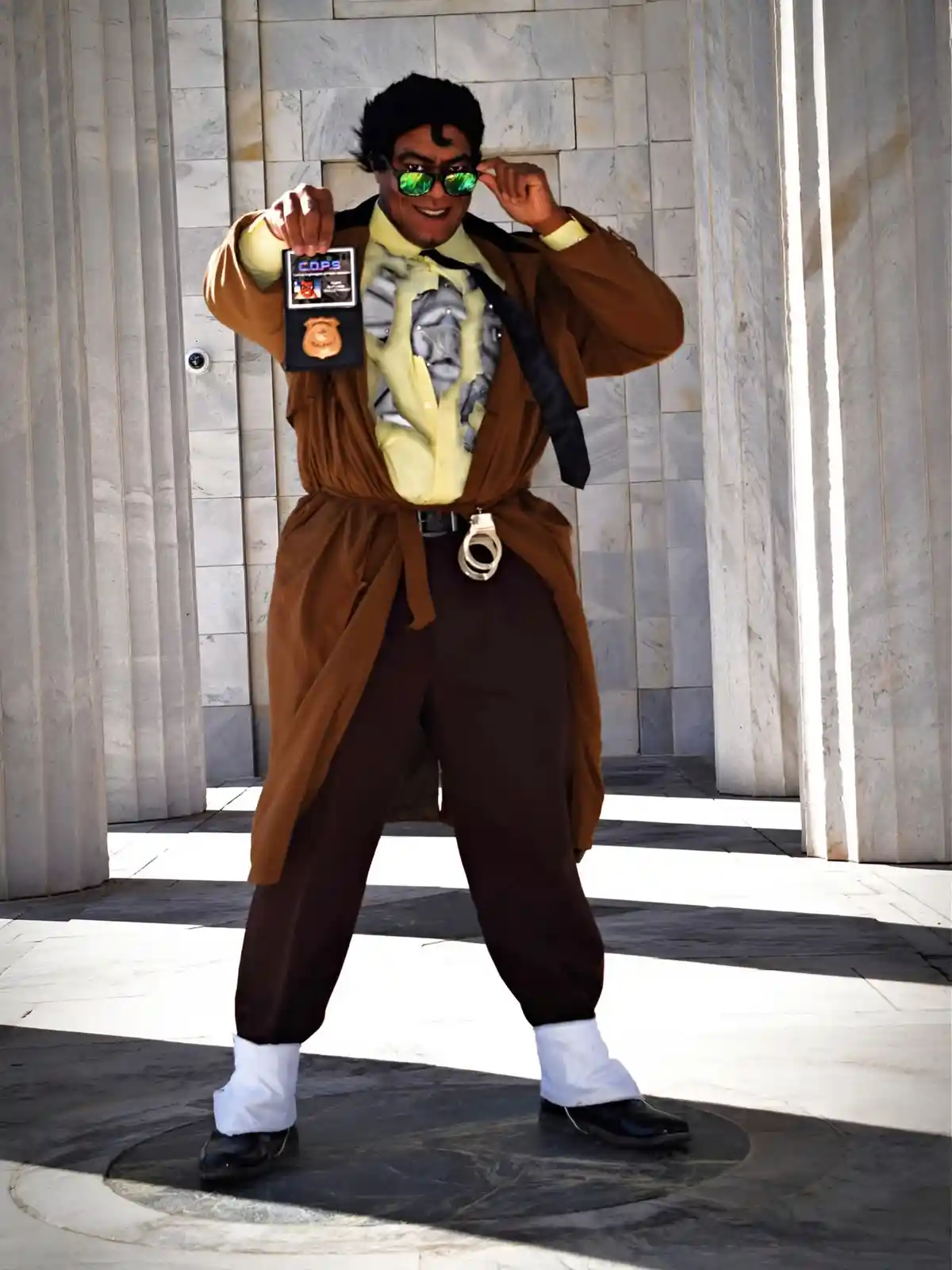 A person dressed in a brown trench coat, yellow shirt, and dark pants, posing confidently with one hand adjusting green…
