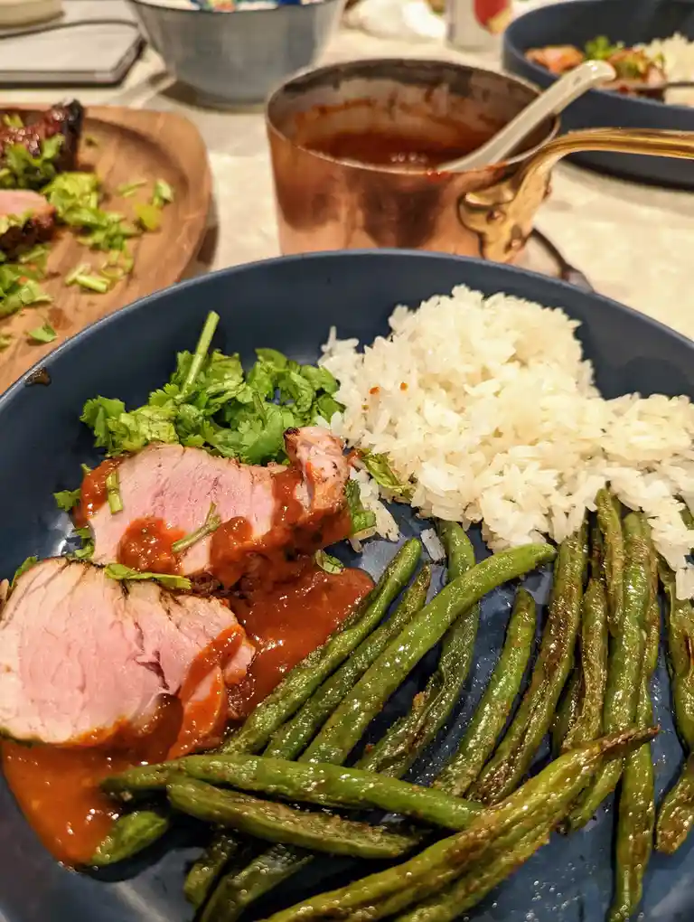 A close-up photo of a blue plate with sliced pork, white rice, and roasted green beans, with a copper saucepan in the…