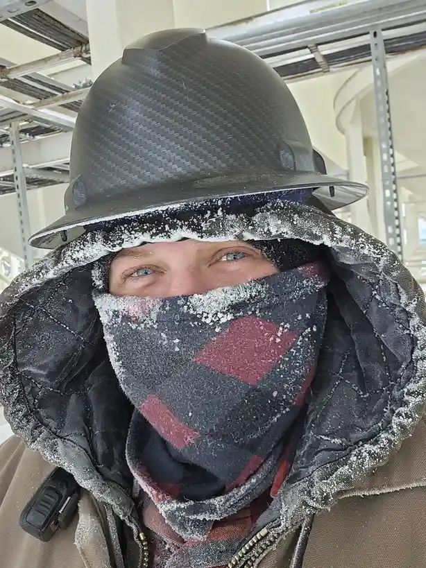 A person wearing a black hard hat and a dark plaid scarf covered in frost, with only their eyes visible, standing in a cold…