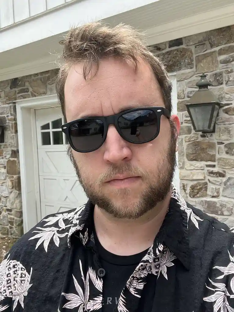A man with a beard and messy hair wears black sunglasses while taking a selfie in front of a stone house with a white door…