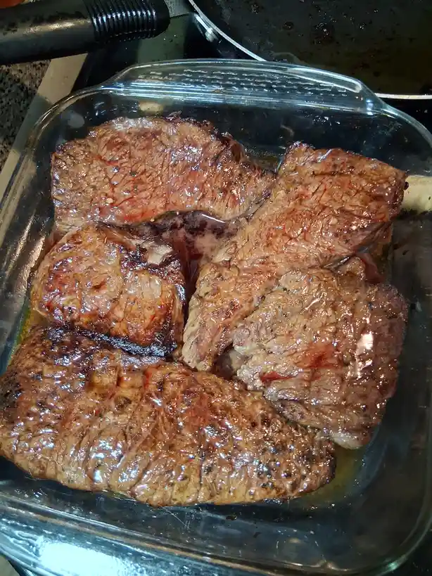 A close-up photo shows several large, seared steaks resting in a clear glass baking dish, glistening with juices and…