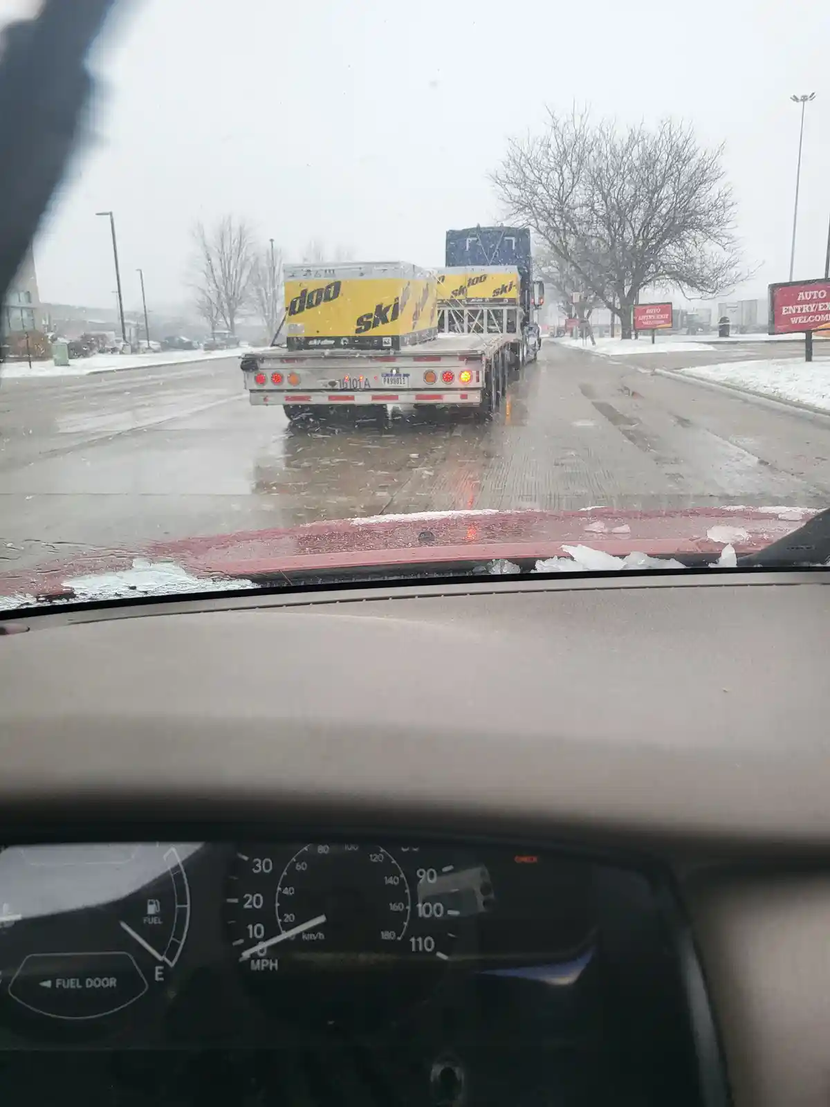 A photo taken from inside a car shows a snowy, wet road with a large flatbed truck ahead carrying two yellow trailers…