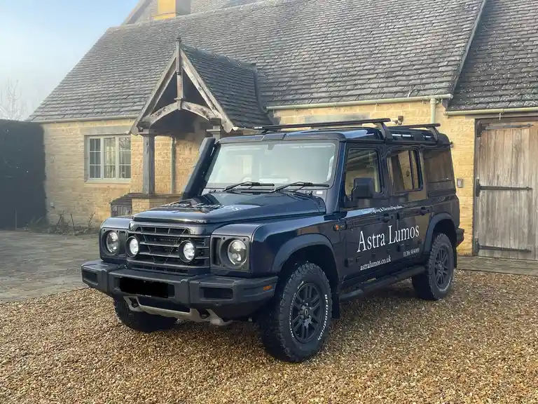 A dark blue, rugged, four-door SUV with 'Astra Lumos' branding and contact details on its side is parked on gravel in front…