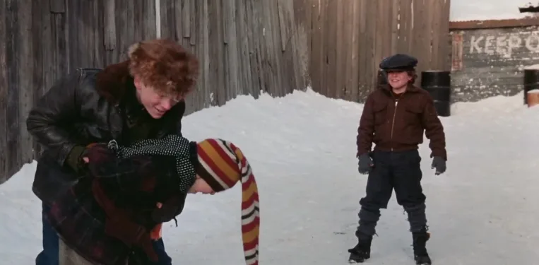 A still from a movie shows two boys in a snowy yard. One boy in a leather jacket and fur hat is holding another boy upside…