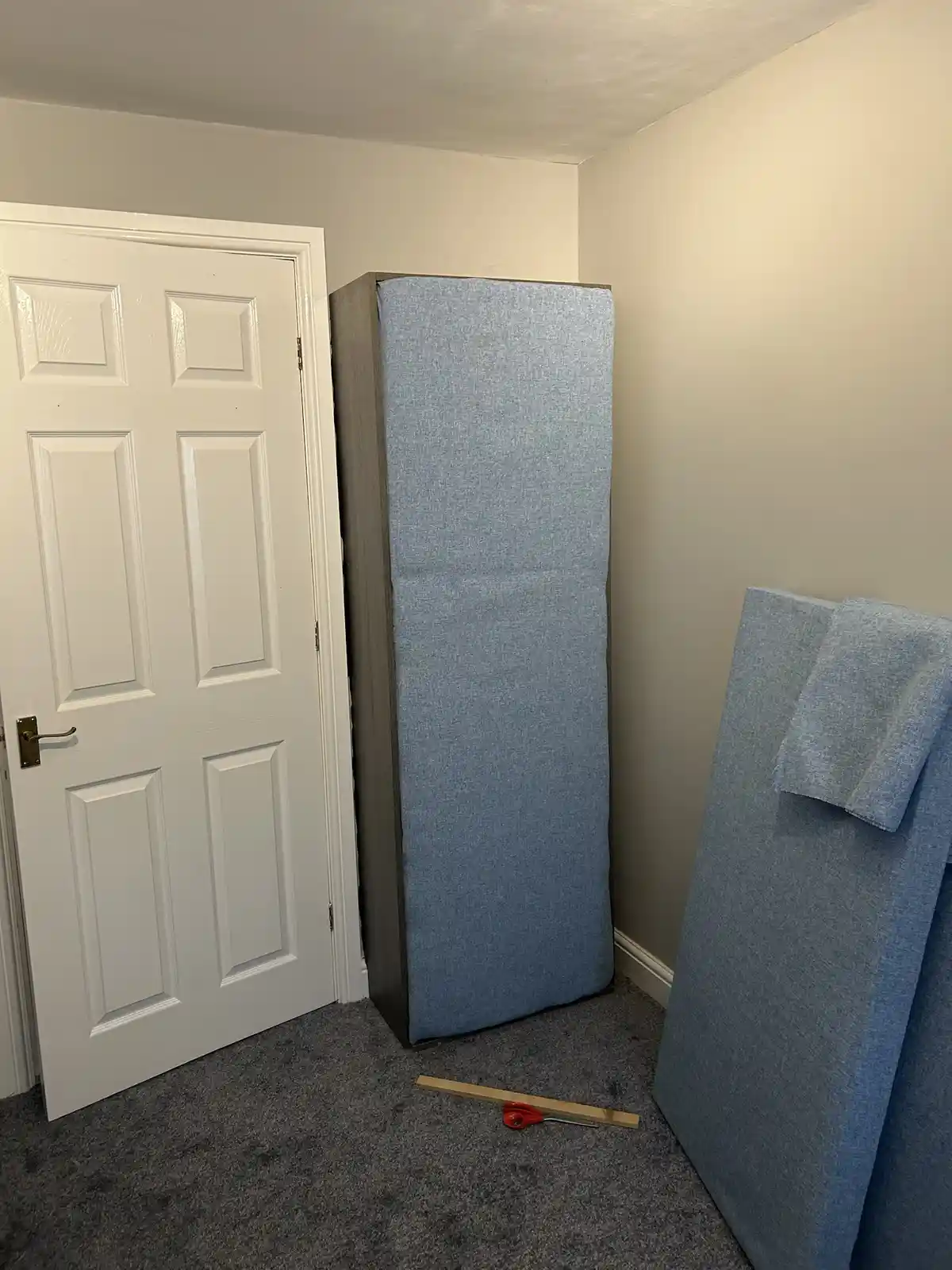 A room with a white door, two large blue upholstered mattresses leaning against walls, and a pair of red-handled scissors…