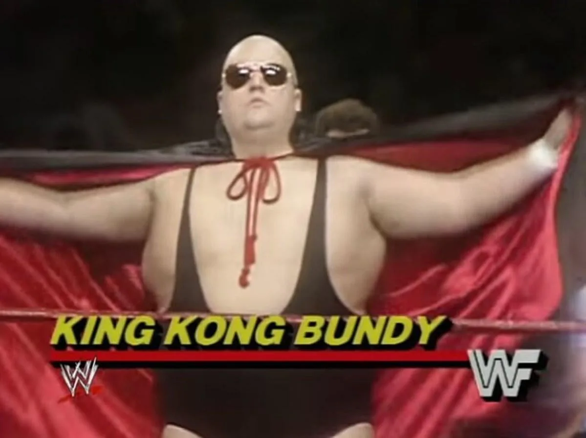 A bald, muscular wrestler wearing sunglasses and a red cape stands in a wrestling ring with arms outstretched. He wears a…