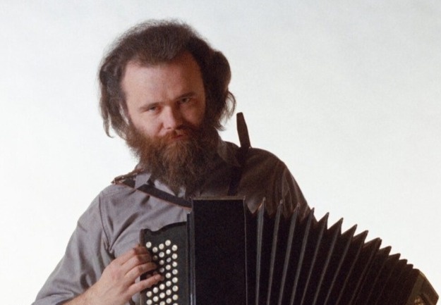 A man with a long beard and dark hair holds a black accordion against a plain white background. He looks directly at the…
