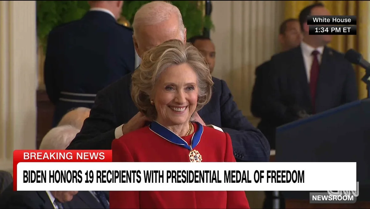 A woman in a red jacket smiles broadly while wearing a Presidential Medal of Freedom around her neck, as a man in a suit…