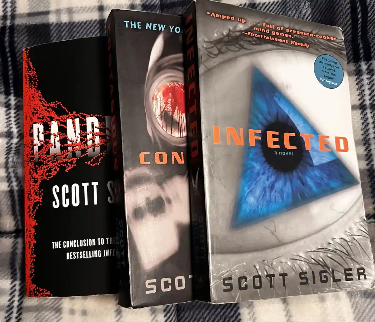 Three books by Scott Sigler are displayed side-by-side on a plaid blanket. The left book, 'Pandemic,' has a black cover with…