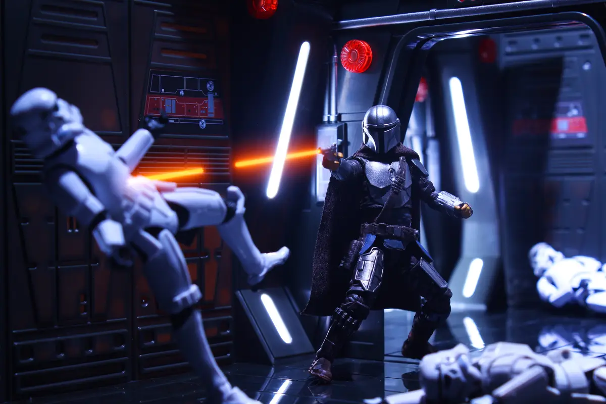 A toy figure of The Mandalorian stands in a sci-fi corridor, firing a blaster at a Stormtrooper action figure who is…
