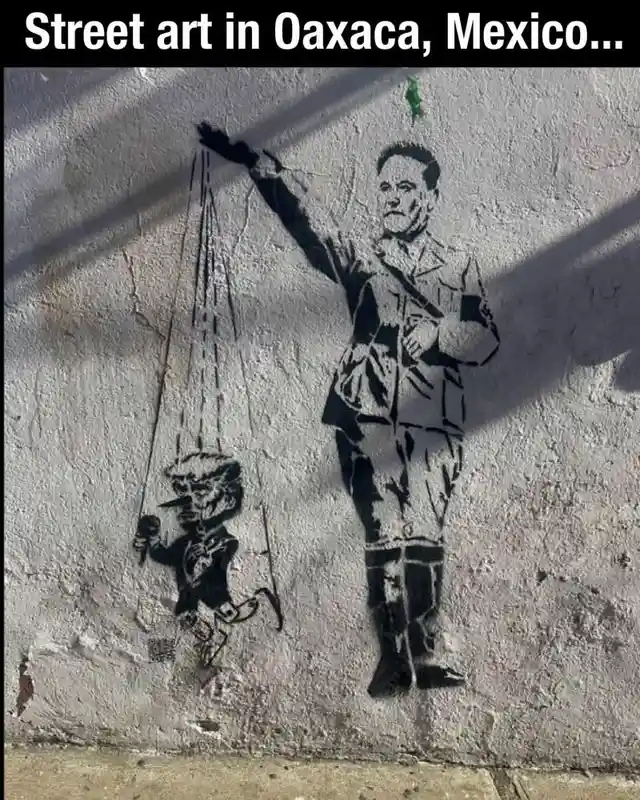 A black-and-white street mural on a weathered wall depicts a large man in a military-style uniform holding puppet strings…