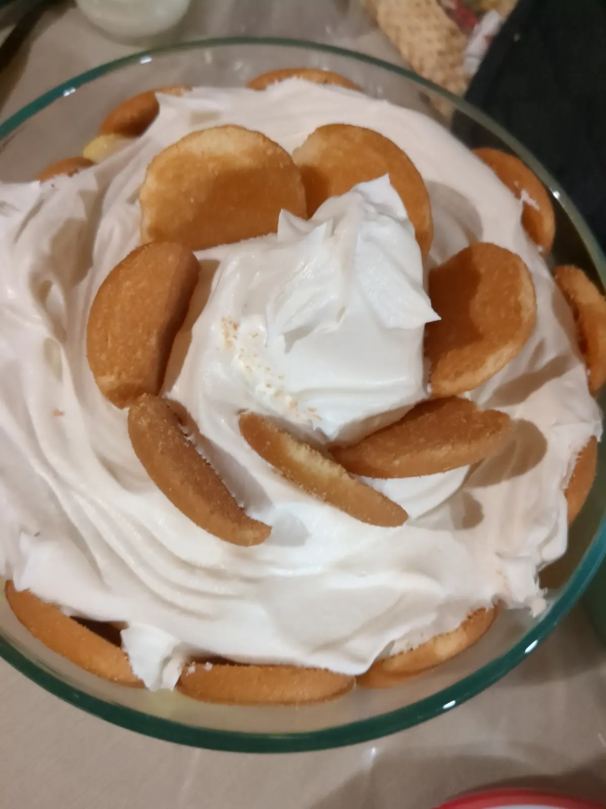 A close-up photo of a dessert in a clear glass bowl, featuring layers of vanilla wafers and a thick white whipped cream…