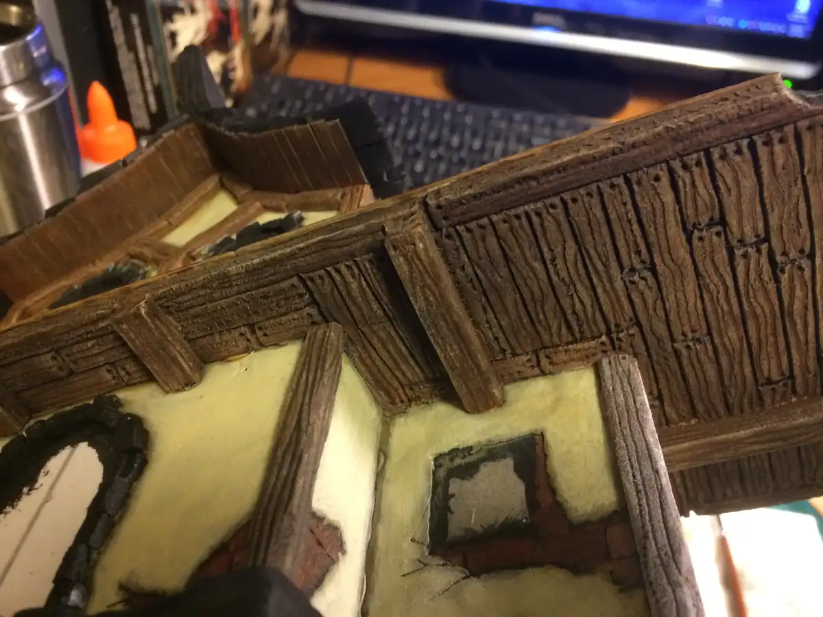 A close-up photo shows a detailed 3d printed miniature building with textured brown wooden roof and walls, painted in beige…
