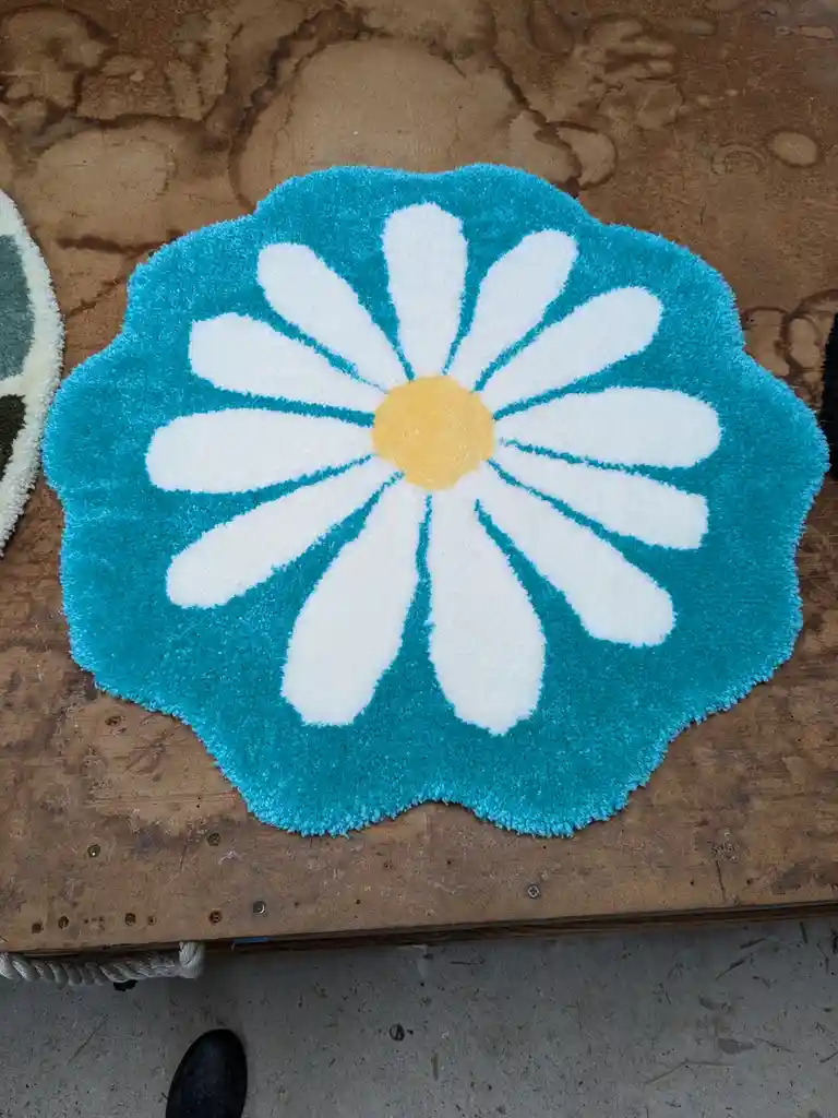 a fluffy turquoise rug with a large white daisy pattern and yellow center sits on a worn wooden surface, with a black shoe…