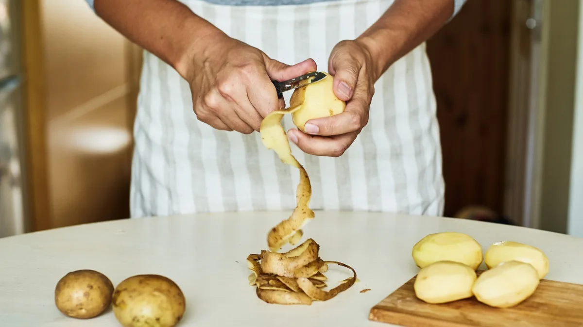 A person wearing a striped apron is peeling a potato with a peeler, with potato peels falling onto a white countertop…