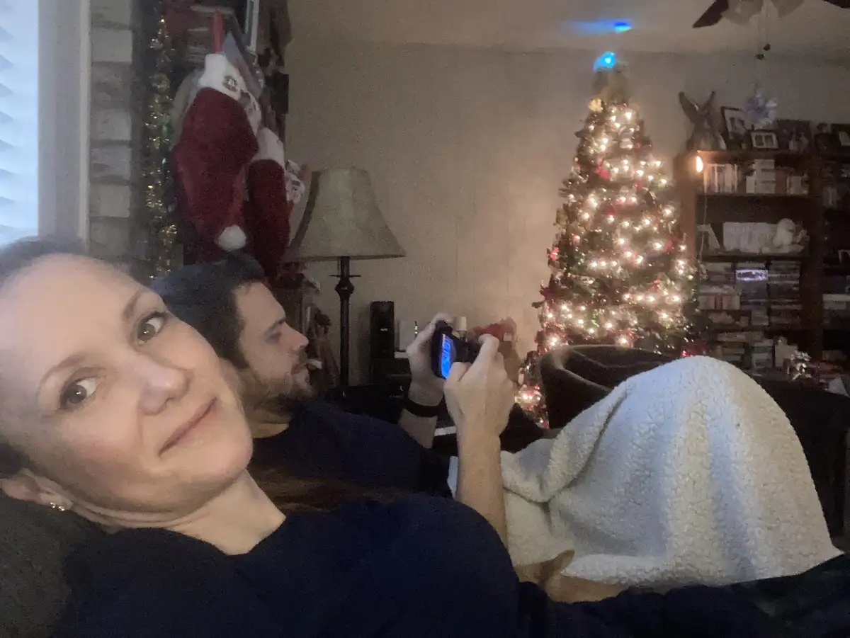 A woman lies on a couch in a living room decorated for Christmas, looking directly at the camera with a tired, slightly…