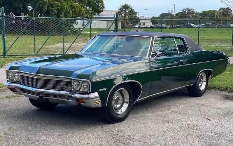 A shiny, dark green classic Chevrolet Chevelle SS 380 from the 1960s is parked on asphalt near a green chain-link fence. The…