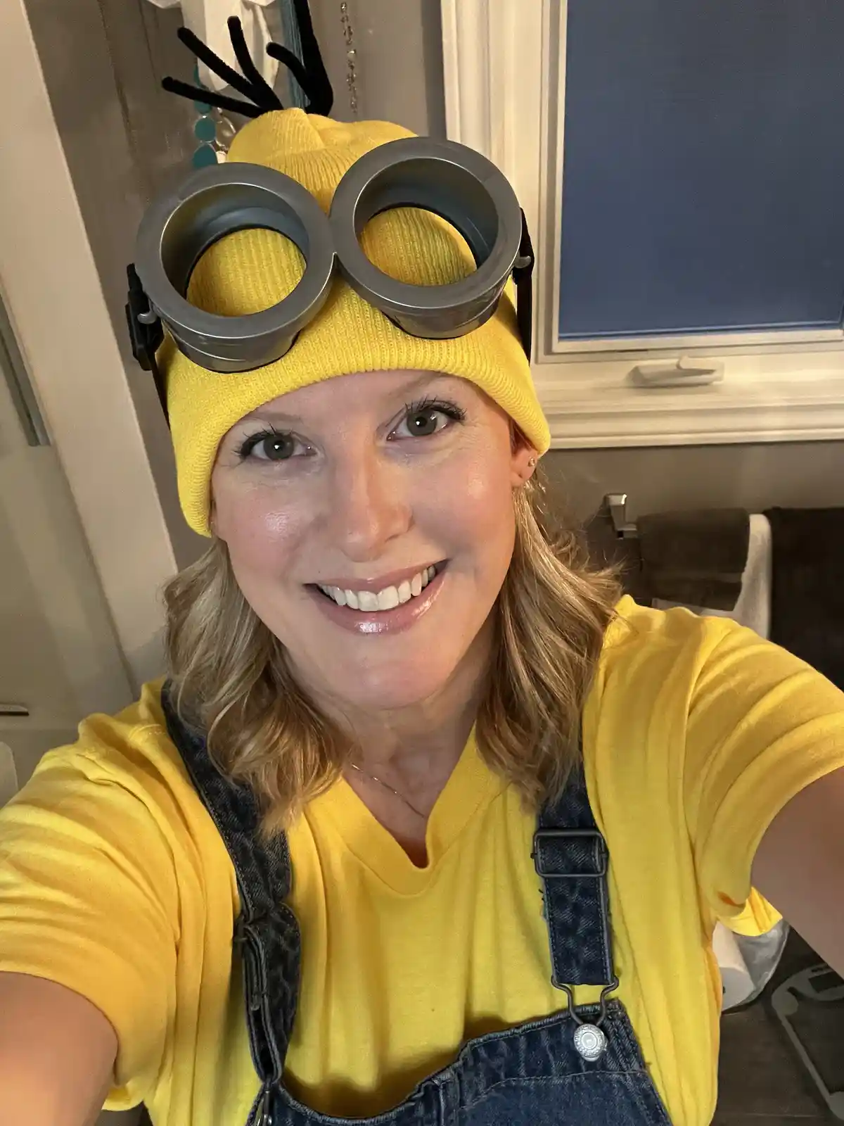 A smiling woman takes a selfie in a bathroom, wearing a yellow beanie with large silver goggles and black antennae, a yellow…