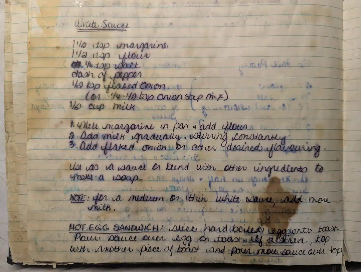 A close-up of a yellowed, lined notebook page with handwritten recipes in blue ink. The top recipe is for 'White Sauce' with…
