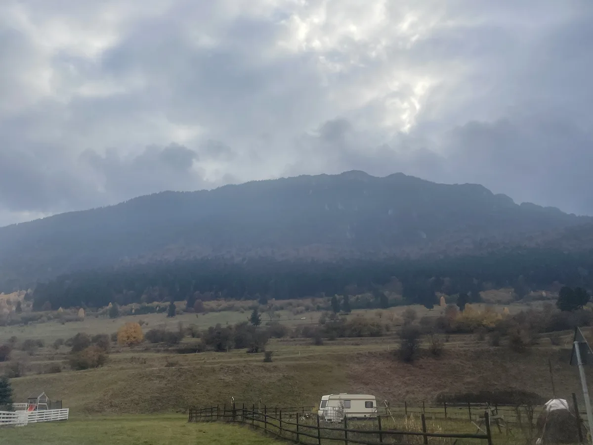 A wide landscape photo shows a cloudy sky over a distant mountain range, with rolling hills, scattered trees showing autumn…