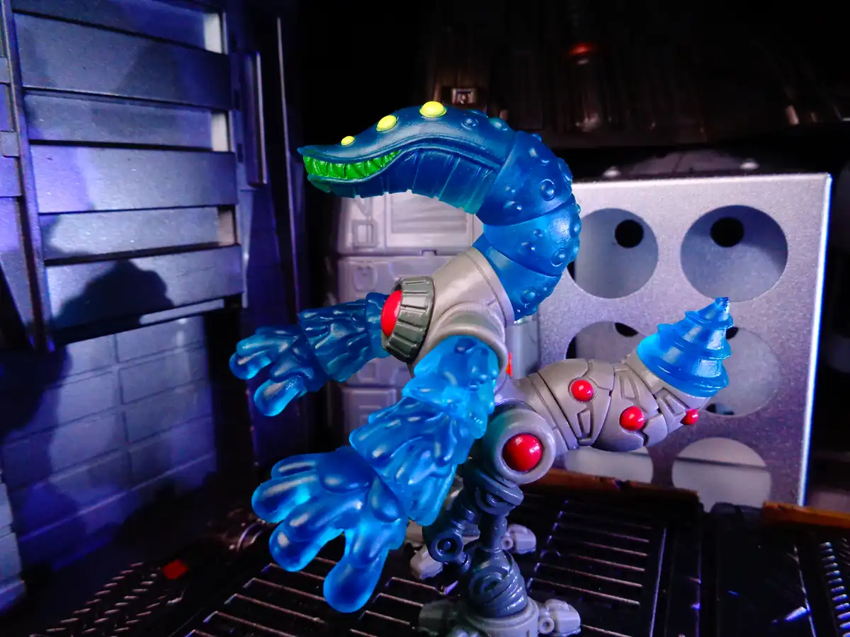 A blue translucent plastic toy figure resembling a robotic crocodile with glowing green teeth and red button eyes stands on…