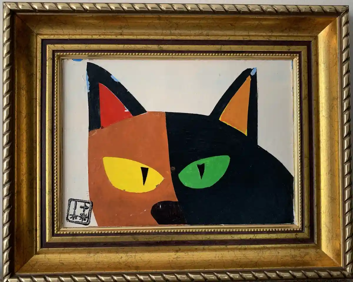 A framed painting depicts a stylized cat split down the middle: one half is orange with a yellow eye, the other half is…