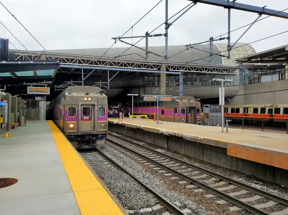 A photo of a train station with multiple trains on adjacent tracks, including a purple and silver train at the front…