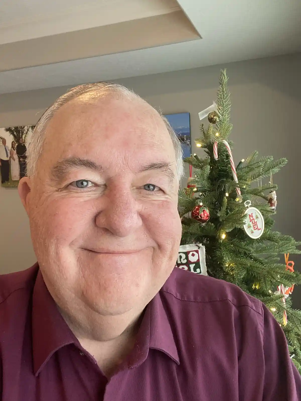 A close-up selfie of a smiling older man with gray hair wearing a maroon collared shirt, standing in front of a decorated…