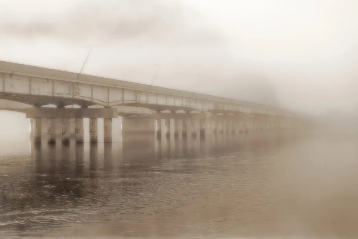 A long, arched concrete bridge stretches over a body of water, partially obscured by thick, hazy fog that blankets the…