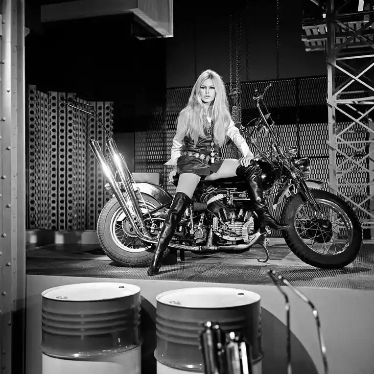 A black-and-white photograph shows a blonde woman in a leather outfit and thigh-high boots sitting on a large motorcycle in…