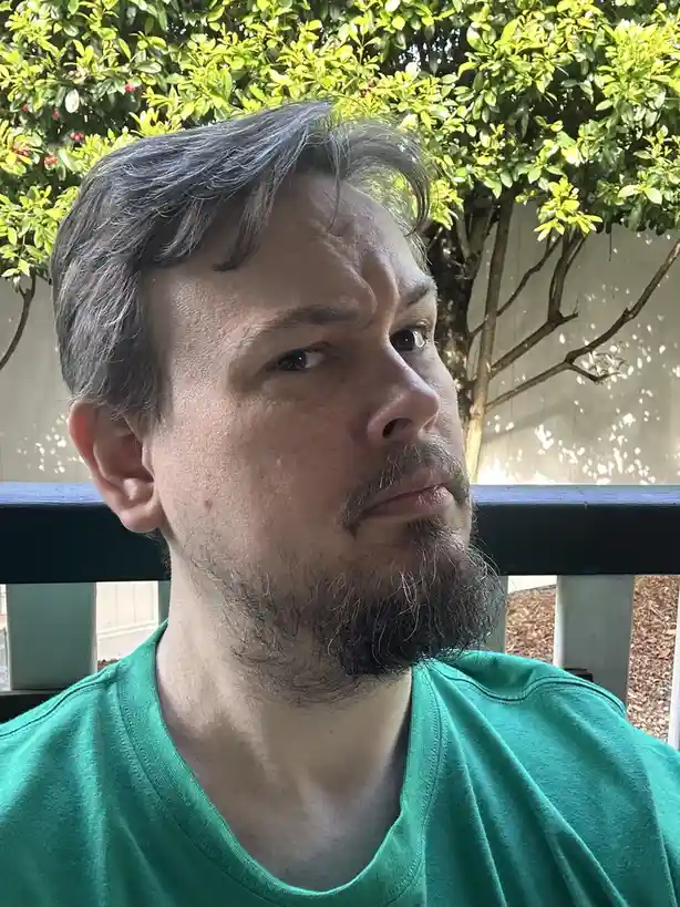 A man with a beard and graying hair, wearing a green t-shirt, looks off to the side with a skeptical, slightly annoyed…