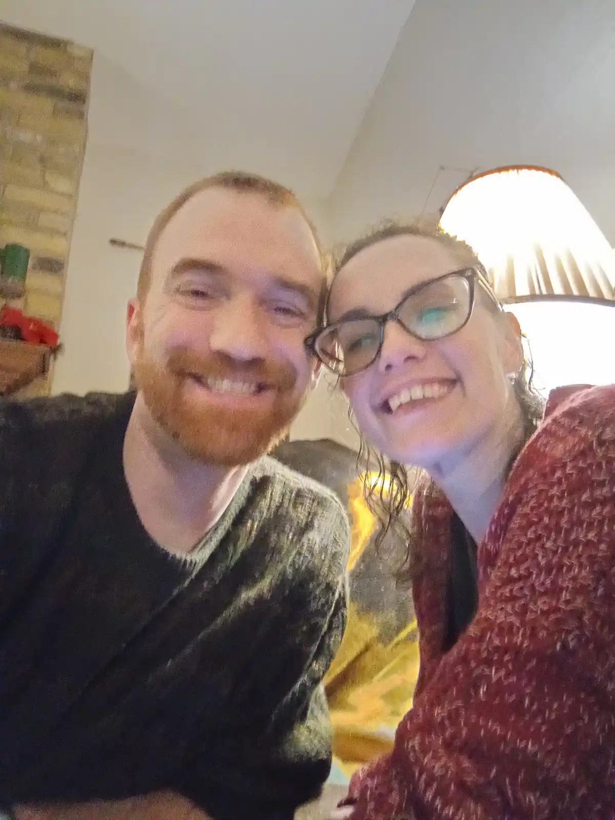 A selfie of a smiling man with a red beard and a smiling woman with glasses, both looking at the camera in a cozy indoor…