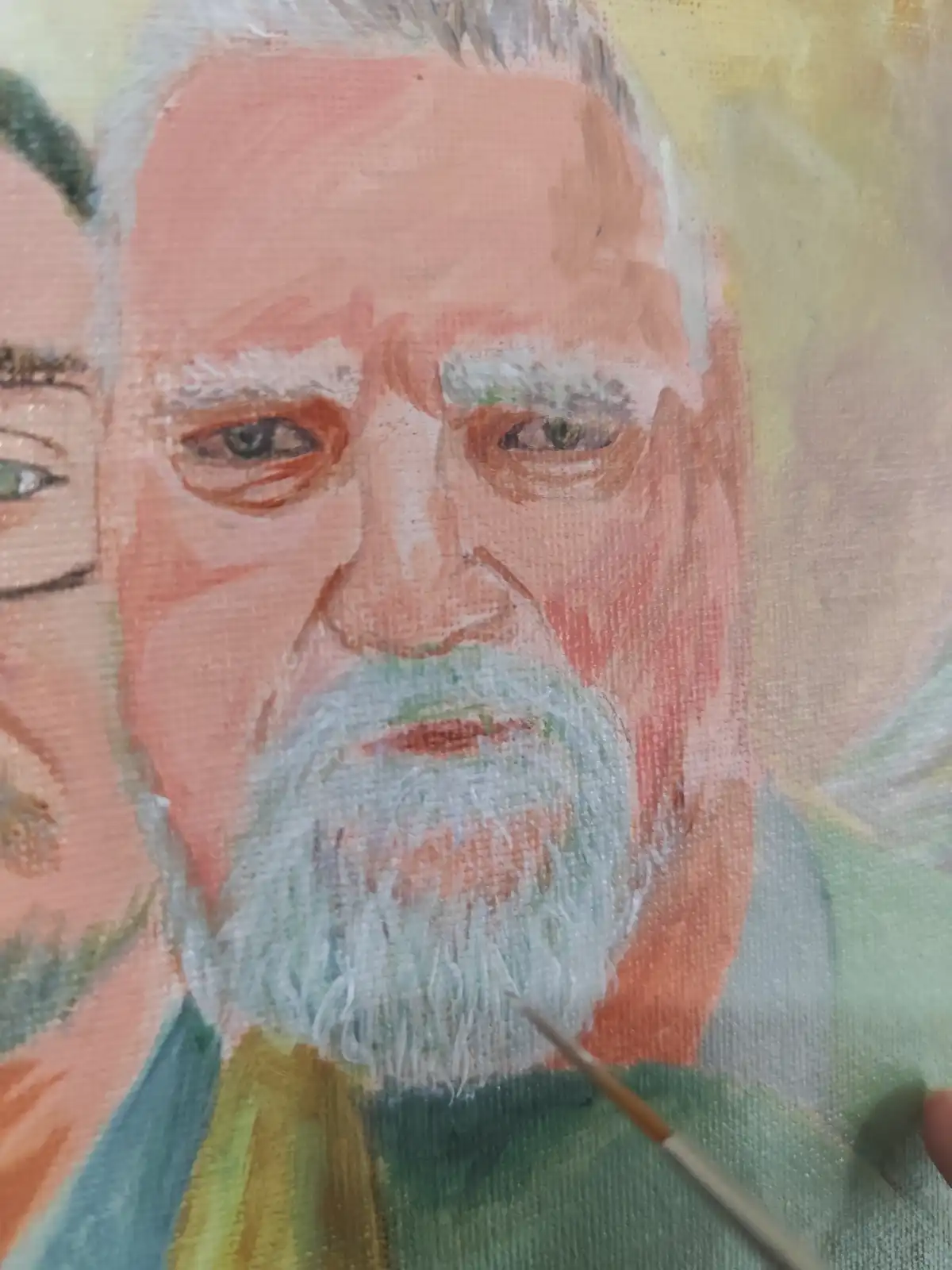 A close-up of an oil painting of an elderly man with a white beard and intense eyes, showing a paintbrush hovering near his…