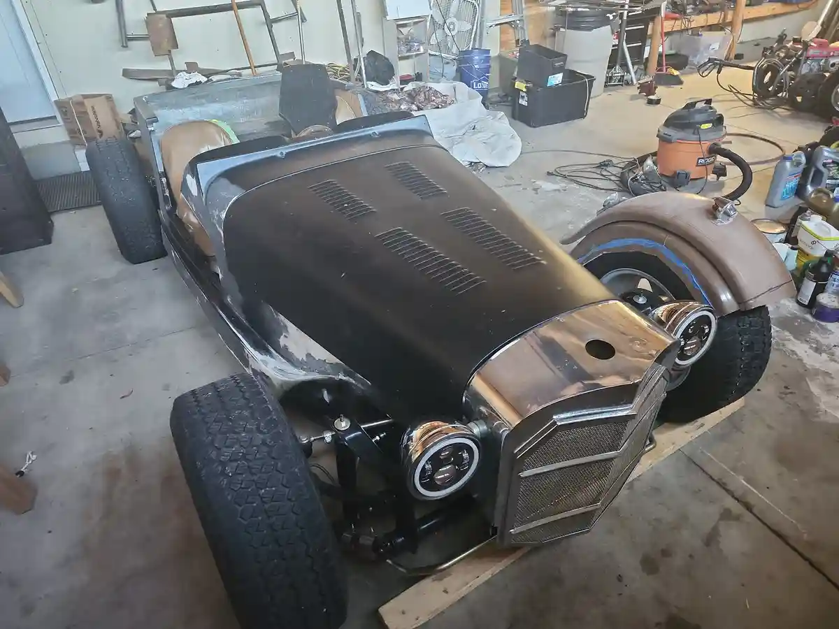 A partially painted vintage-style custom car sits in a cluttered garage, its body mostly black with some unpainted or white…