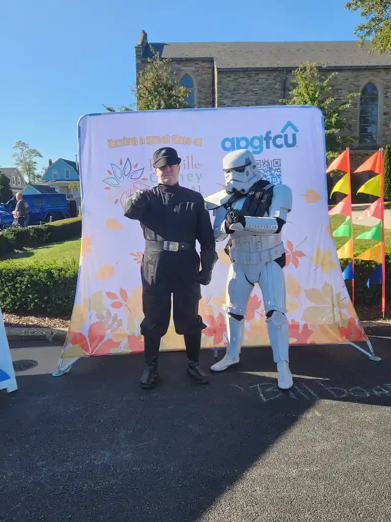Two people pose in front of a festival backdrop: one dressed as a stormtrooper and the other in a black uniform resembling a…