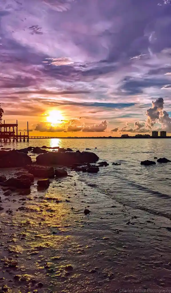 A scenic sunset photo shows a vibrant purple and orange sky over water with rocks in the foreground and a pier on the left…