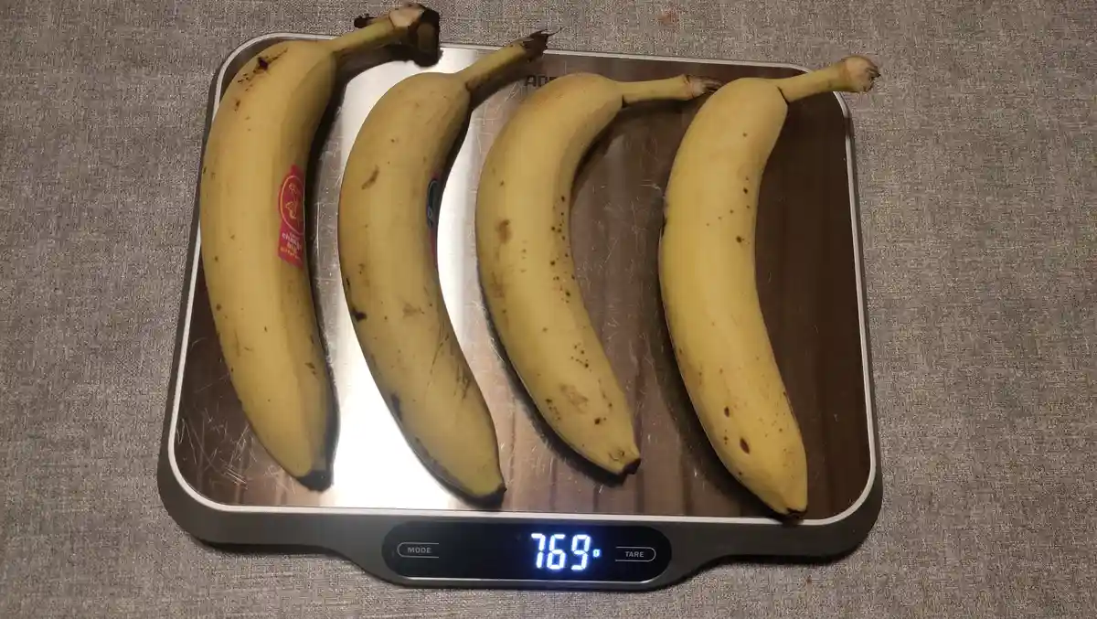 Four ripe yellow bananas with brown spots sit on a digital kitchen scale, which displays the weight as 769g. The bananas are…