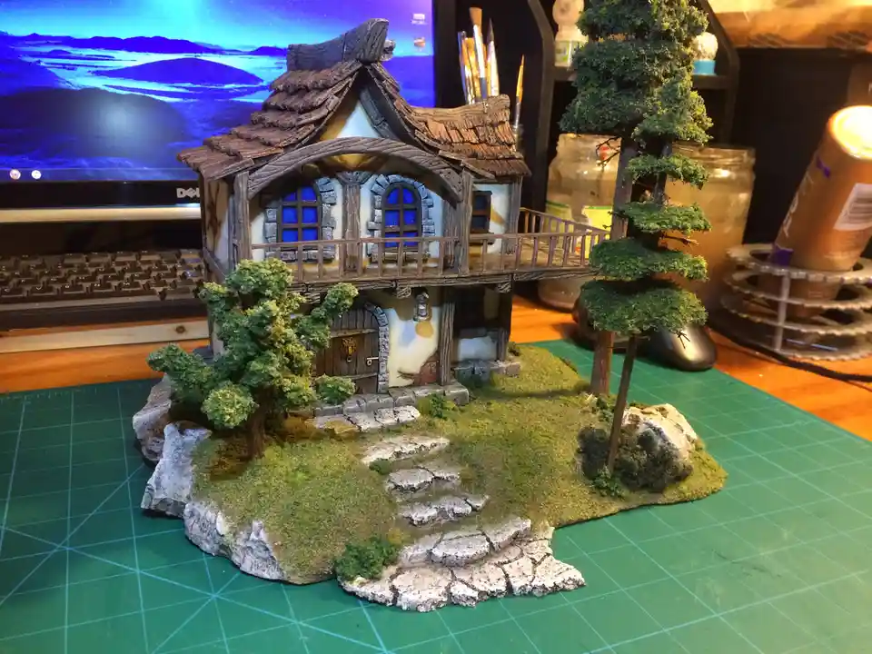 A highly detailed miniature fantasy cottage with a wooden roof, stone foundation, and blue windows sits on a green cutting…