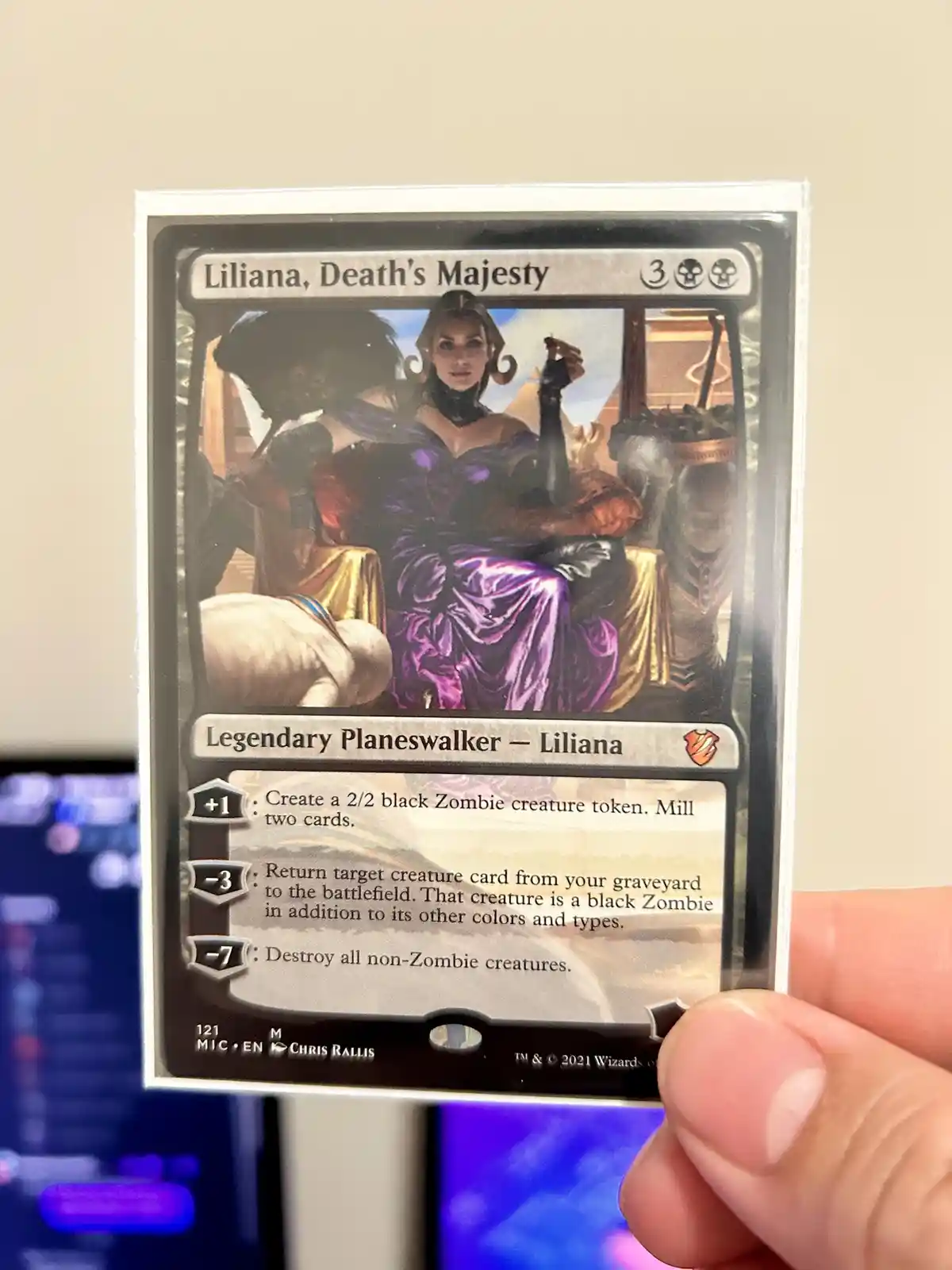 A hand holds a Magic: The Gathering card titled 'Liliana, Death's Majesty,' showing a woman in a purple gown seated on a…