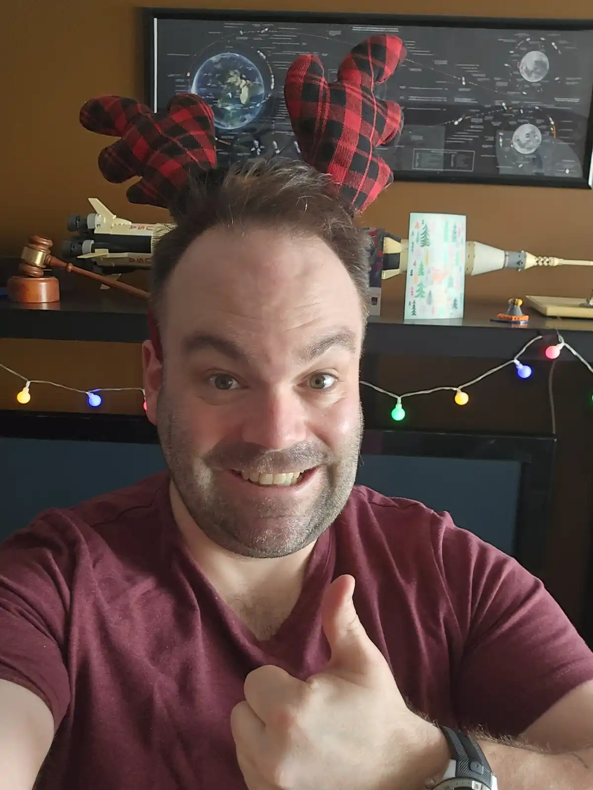 A man with a beard and short brown hair wears red-and-black plaid reindeer antlers and gives a thumbs-up while smiling at…