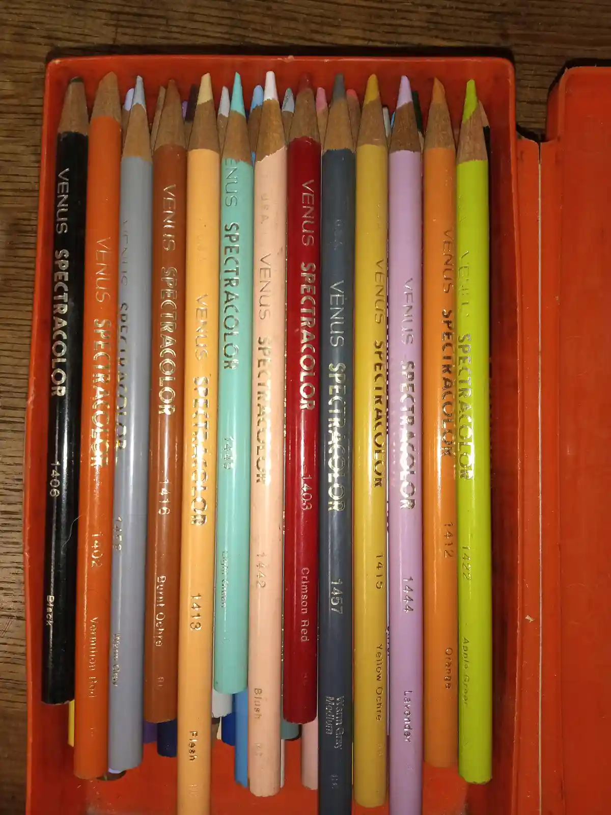 A close-up photo shows a row of used Venus Spectracolor colored pencils arranged in an orange plastic case, with their…