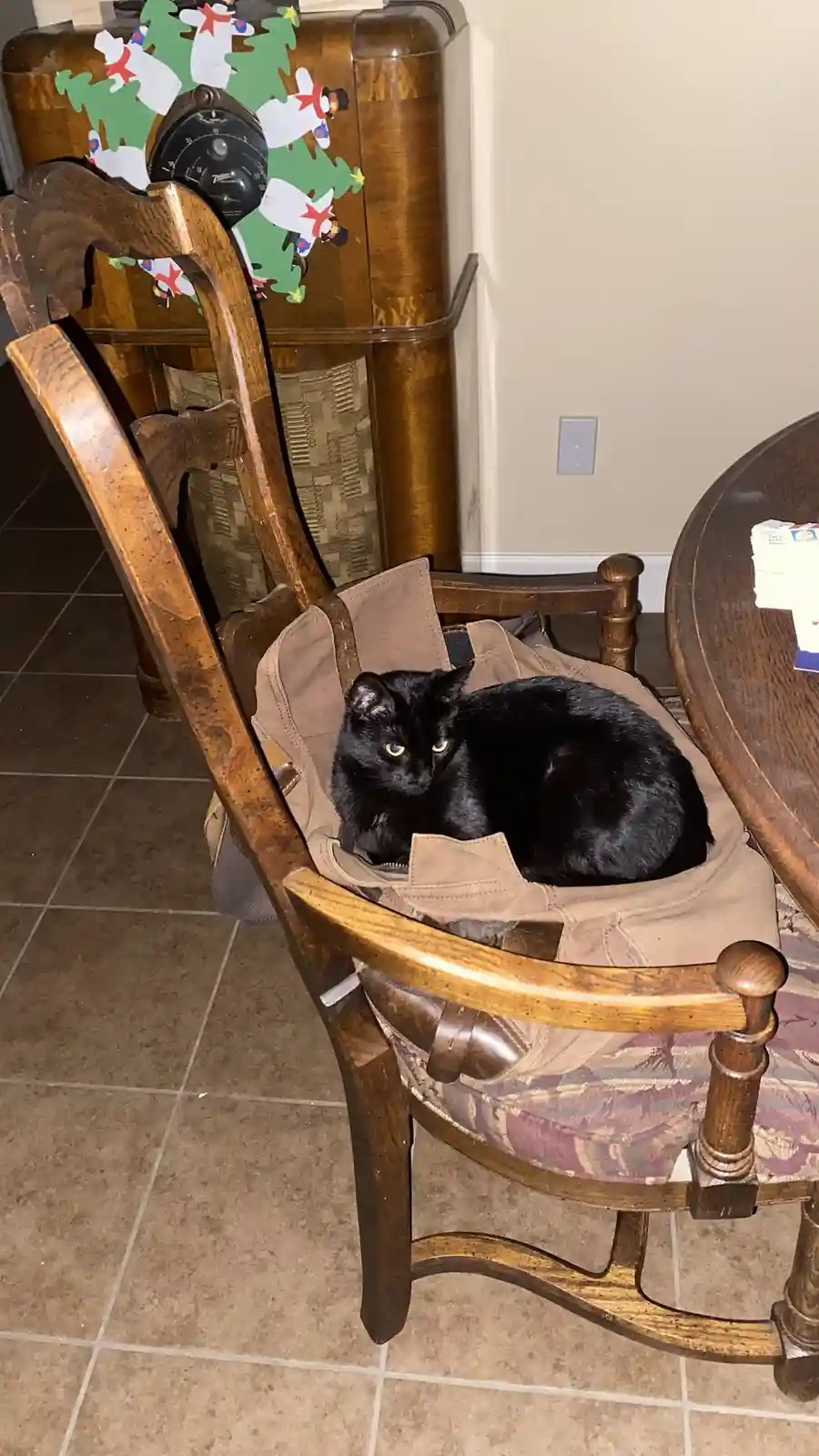 A black cat with yellow eyes is curled up and resting inside a tan jacket that is draped over the seat of a wooden rocking…
