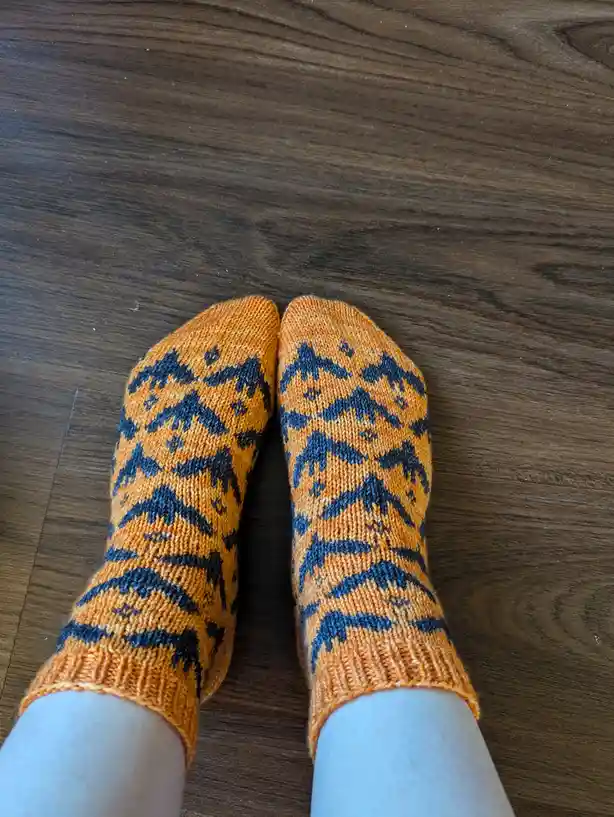 A top-down photo shows a person's feet wearing orange and dark blue knitted socks with a repeating pattern of stylized…